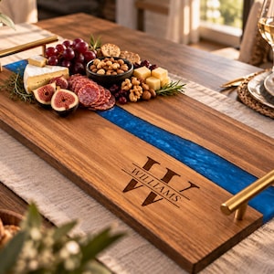 May include: A rectangular wooden serving tray featuring a central blue resin river inlay, gold-toned metal handles, and the engraved text "WILLIAMS" with a decorative monogram. The tray is arranged with assorted cheeses, grapes, figs, and nuts.