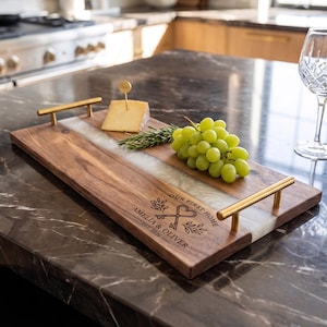 May include: A rectangular wooden serving tray with a central white resin inlay and two gold-toned metal handles. The wood is engraved with the text "OUR FIRST HOME", "AMELIA & OLIVER", and "EST. 2026" alongside a graphic of two keys and leaves.