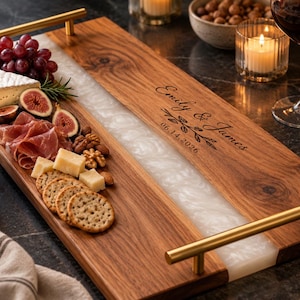 May include: A rectangular wooden serving tray with a central white resin inlay, gold-toned metal handles, and the engraved text Emily & James 06.14.2026. The tray is arranged with grapes, cheese, figs, prosciutto, nuts, and crackers.