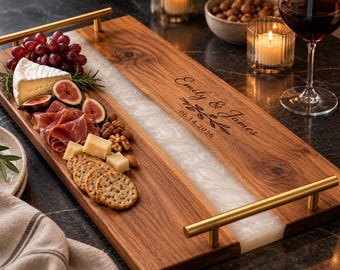 May include: A rectangular wooden serving tray with a central white resin inlay, gold-toned metal handles, and the engraved text Emily & James 06.14.2026. The tray is arranged with grapes, cheese, figs, prosciutto, nuts, and crackers.