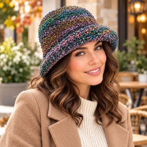 Può includere: Un cappello a secchiello caratterizzato da un motivo a maglia testurizzato e multicolore con tonalità vivaci di verde, viola, blu e oro. Il cappello presenta una corona arrotondata e una tesa spiovente, realizzato in filato spesso e variegato.