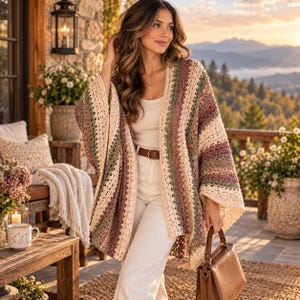 May include: A crochet cardigan featuring vertical stripes in cream, sage green, and muted mauve. The garment has an open front, wide sleeves, and a textured, lacy stitch pattern. It is worn over a cream-colored top and white trousers with a brown belt.