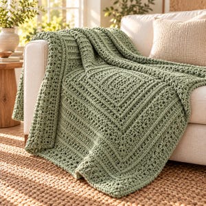 Puede incluir: Una manta de ganchillo color verde salvia con un patrón cuadrado texturizado que alterna filas de puntos densos y detalles calados. La manta está colocada sobre el brazo de un sofá, mostrando su construcción de hilo grueso y color uniforme.