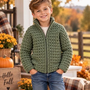 May include: A sage green long-sleeve zip-up sweater with a textured waffle-knit pattern, a high collar, and a dark zipper closure, worn over a plain white shirt paired with blue denim jeans, showcasing a classic casual style for cooler weather.