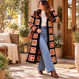 May include: A long, open-front crochet coat featuring a repeating granny square pattern in shades of black, white, orange, and red. The coat is worn over a white mock-neck top and light blue denim jeans, paired with brown leather boots.