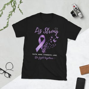 May include: A black short-sleeve t-shirt featuring a purple awareness ribbon and a swarm of purple butterflies. The text reads: Liz Strong, FAITH. HOPE. STRENGTH. LOVE. We fight together, accompanied by two small heart outlines in purple.