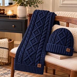 Pode incluir: Um conjunto de cachecol e gorro de malha em azul-marinho profundo, com um padrão complexo de tranças e formas de diamante. Ambas as peças incluem uma pequena etiqueta quadrada de couro castanho-claro com um ícone de árvore.