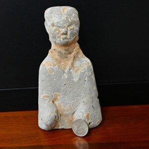 May include: A weathered, light gray stone figurine of a person with a smooth, rounded head and a simple, block-like torso. The surface shows patches of tan discoloration and texture. A silver-colored United States quarter coin is placed at the base.