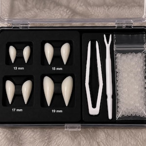 May include: A black storage case containing four pairs of white vampire fangs in sizes 13 mm, 15 mm, 17 mm, and 19 mm. The set includes a pair of white plastic tweezers, a white applicator tool, and a small clear bag filled with adhesive beads.