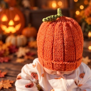 May include: A bright orange knitted beanie hat designed to resemble a pumpkin, featuring vertical ribbed texture and a small green crocheted stem on top. The hat is worn over a white long-sleeved garment decorated with a repeating pattern of small orange pumpkins.