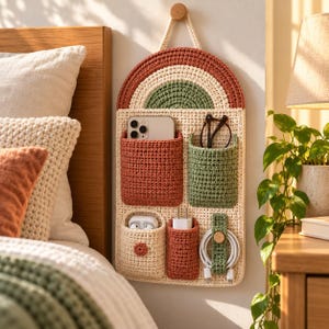 Puede incluir: Un organizador de pared de ganchillo con base color crema y una parte superior de arcoíris semicircular en tonos óxido y verde salvia. Incluye cinco bolsillos de varios tamaños que contienen un teléfono inteligente, gafas, auriculares y un cable.