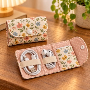 May include: A floral print fabric organizer case with a snap button closure. The exterior features a cream background with pink, yellow, and blue flowers. The interior is lined with pink polka dot fabric and includes elastic loops for cables.