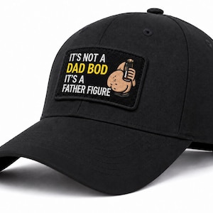 May include: A black baseball cap featuring a rectangular patch on the front. The patch displays the text IT'S NOT A DAD BOD IT'S A FATHER FIGURE in white and yellow lettering, alongside a cartoon illustration of a round, tan-colored character.