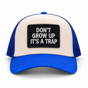 May include: A blue and cream trucker cap featuring a rectangular black patch on the front panel. The patch displays the white embroidered text DON'T GROW UP IT'S A TRAP. The cap has a solid blue brim, a cream-coloured front, and blue mesh back panels.
