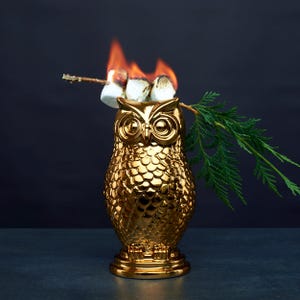 May include: A metallic gold owl-shaped container with a textured feather pattern. A wooden skewer rests across the top, holding three toasted marshmallows with visible flames, accompanied by a small green evergreen branch extending to the right.