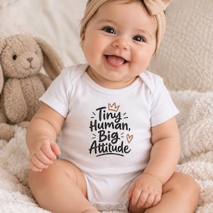 Op de afbeelding: Een witte babyromper met korte mouwen en de zwarte tekst Tiny Human, Big Attitude, voorzien van een klein bruin kroontje en hartje. Het kledingstuk heeft een ronde halslijn en drukknopen aan de onderzijde voor baby's om te dragen.