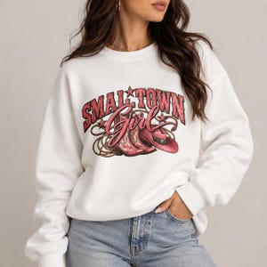 Op de afbeelding: Een wit sweatshirt met lange mouwen en een grafische print met de tekst "SMALL TOWN Girl" in rood en roze. Het ontwerp bevat een paar roze cowboylaarzen, een roze cowboyhoed en een opgerolde lasso met kleine steraccenten.
