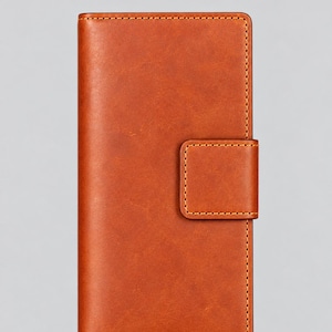 May include: A rectangular, tan-coloured wallet with a smooth, matte finish and visible stitching along the edges. The wallet features a vertical orientation and a small, rectangular flap closure on the right side, also secured with matching stitching.