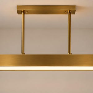Könnte beinhalten: Eine lineare Pendelleuchte mit gebürstetem Messing-Finish. Der rechteckige Rahmen verfügt über zwei vertikale Stangen, die mit einer horizontalen Deckenhalterung verbunden sind. Die Unterseite hat eine flache, weiße Lichtdiffusionsplatte.