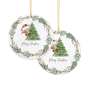 Könnte beinhalten: Zwei runde, weiße Keramikornamente, die an goldenen Schnüren hängen. Jedes zeigt eine Aquarellillustration eines braunen Teddybären mit roter Weihnachtsmütze hinter einem grünen Tannenbaum, umrahmt von einem Tannenkranz und dem Schriftzug Merry Christmas.