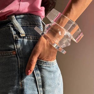 May include: A set of two clear, transparent acrylic bangles with a geometric, faceted design. The bracelets feature a smooth, polished surface that catches the light, creating subtle reflections and a high-gloss appearance on the wrist.