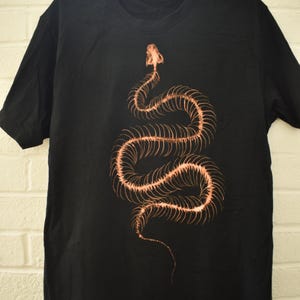 Peut inclure: Un t-shirt noir à manches courtes avec une impression graphique couleur cuivre représentant un squelette de serpent. Le motif squelettique montre le crâne et la colonne vertébrale sinueuse du serpent centré sur le devant du vêtement.