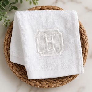 May include: A white terry cloth hand towel featuring a centered, tone-on-tone embroidered monogram of the letter H. The letter is enclosed within a decorative, square-shaped frame with rounded corners, showcasing a textured, woven stitch pattern.