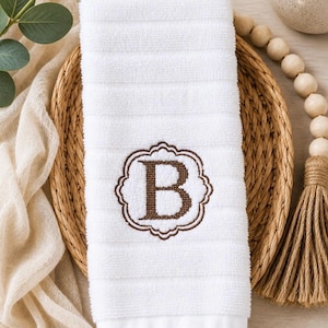 May include: A white hand towel featuring a textured, ribbed surface and a centered brown embroidered monogram of the letter B, enclosed within a decorative scalloped frame. The towel is rectangular with a smooth, flat hem at the bottom edge.