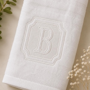 Può includere: Un asciugamano da bagno in cotone bianco con un bordo testurizzato e un monogramma centrale ricamato della lettera B. La lettera è racchiusa all'interno di una cornice rettangolare decorativa e smerlata, realizzata in filo bianco.
