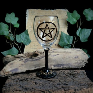Può includere: Un bicchiere di vino nero con un design a pentagramma dorato. Il bicchiere è appoggiato su un pezzo di legno alla deriva.