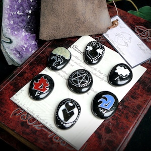 Op de afbeelding: Set van acht zwarte stenen met verschillende symbolen erop geschilderd. De stenen zijn gerangschikt op een bruin lederen gebonden boek met de tekst "Stones" en "Dam Crafted" gedrukt op de pagina. De stenen vertegenwoordigen de vier elementen: aarde, lucht, vuur en water.