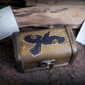 Può includere: Un piccolo forziere in legno con coperchio incernierato e chiusura in metallo. Il forziere è dipinto con un disegno di corvo nero e stelle dorate. Il testo "Raven" è visibile su una carta sullo sfondo. Il forziere viene probabilmente utilizzato per riporre piccoli oggetti.