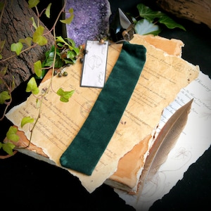 May include: A green velvet bookmark with a decorative tassel, lying on top of an old, worn book with handwritten text. A feather pen and a purple crystal are also visible in the image.