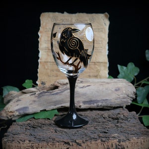 May include: A clear wine glass with a black stem, decorated with a hand-painted black raven and gold details. The raven sits on a branch with a white moon. The background includes vintage paper and wood.