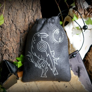 Peut inclure: Une pochette en velours noir avec un motif brodé blanc représentant un corbeau perché sur une branche avec un motif de lune et de spirale en arrière-plan. La pochette est munie d'un cordon de serrage et est parfaite pour ranger de petits objets.