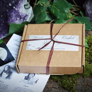 May include: A brown cardboard box with a red ribbon tied around it. The box has a label that says "Hand Crafted" and "Moon Goddess Garden.com".