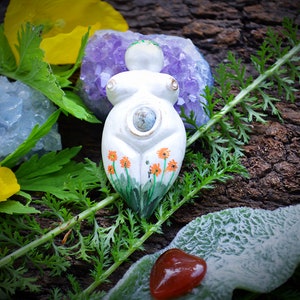 May include: A white ceramic goddess figurine with orange flower details, surrounded by greenery, crystals, and a red stone. The figurine has a blue-green center and is set against a dark, textured background. Yellow flowers and blue crystals add color.
