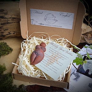 May include: A small, hand-crafted terracotta figurine of a fertility goddess with a spiral design. The figurine is in a small box with shredded paper and a card that reads "Fertility Spiral Goddess".