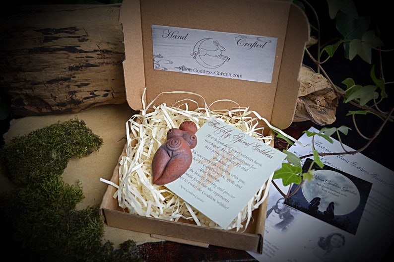 May include: A small, hand-crafted terracotta figurine of a fertility goddess with a spiral design. The figurine is in a small box with shredded paper and a card that reads "Fertility Spiral Goddess".