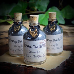May include: Three small glass bottles with cork stoppers filled with black salt. Each bottle has a black label with gold lettering that reads "Witches Black Salt - Drive away Evil - Hand Blended by Moon Goddess Garden".
