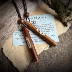 K&ouml;nnte beinhalten: Zwei Holz-Talismane mit geschnitzten Symbolen, an braunen Kordeln aufgeh&auml;ngt. Einer ist ein dunkleres Rotbraun, der andere ein helleres Hellbraun. Im Hintergrund befindet sich eine kleine Karte mit Text &uuml;ber die Talismane. Die Talismane sind ca. 10 cm lang.