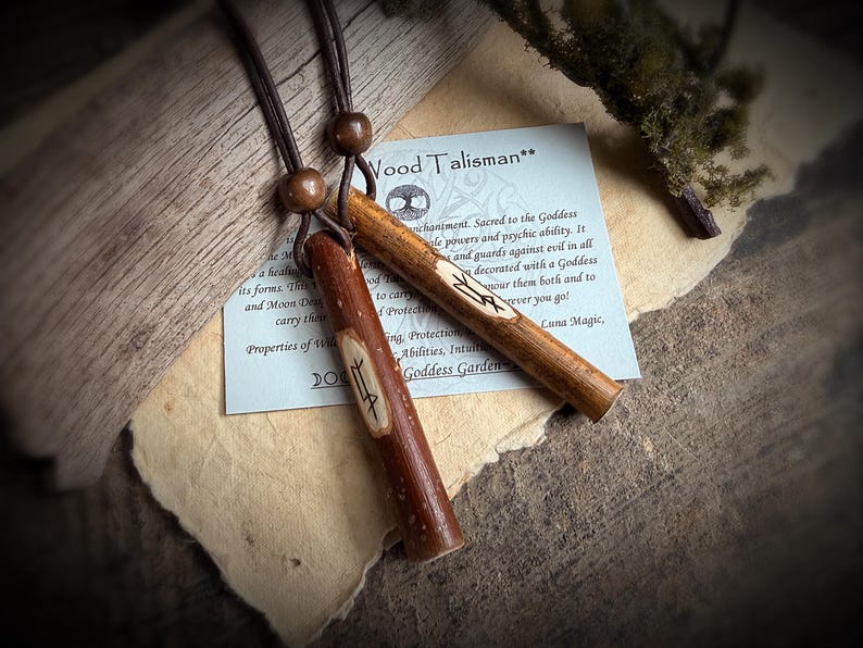 K&ouml;nnte beinhalten: Zwei Holz-Talismane mit geschnitzten Symbolen, an braunen Kordeln aufgeh&auml;ngt. Einer ist ein dunkleres Rotbraun, der andere ein helleres Hellbraun. Im Hintergrund befindet sich eine kleine Karte mit Text &uuml;ber die Talismane. Die Talismane sind ca. 10 cm lang.