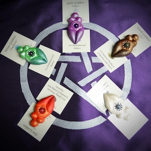 May include: Five ceramic goddess figurines, each representing a different element: water, fire, earth, spirit, and air. Each figurine is hand-painted and has a unique design. The figurines are displayed on a purple cloth with a silver pentagram design.
