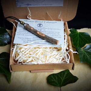 May include: A Rowan Wood Talisman necklace in a cardboard box with shredded paper. The wooden pendant has carved symbols and a leather cord. The box is surrounded by green leaves and berries.