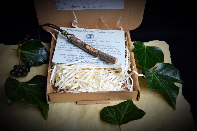 May include: A Rowan Wood Talisman necklace in a cardboard box with shredded paper. The wooden pendant has carved symbols and a leather cord. The box is surrounded by green leaves and berries.