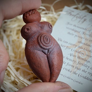 May include: A small, terracotta figurine of a fertility goddess with a spiral design on her belly. The figurine is being held in a hand, and the background is a brown paper with shredded paper.