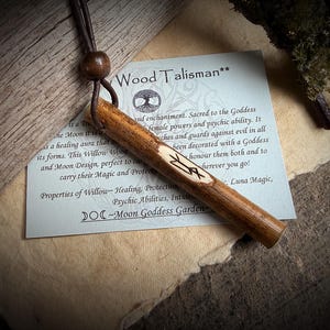 K&ouml;nnte beinhalten: Ein Holz-Talisman mit einer Lederschnur und einer Holzperle. Der Talisman ist zylindrisch, mit einem geschnitzten Symbol. Eine Karte mit Text &uuml;ber die Eigenschaften des Talismans liegt daneben. Der Text auf der Karte lautet "Wood Talisman".
