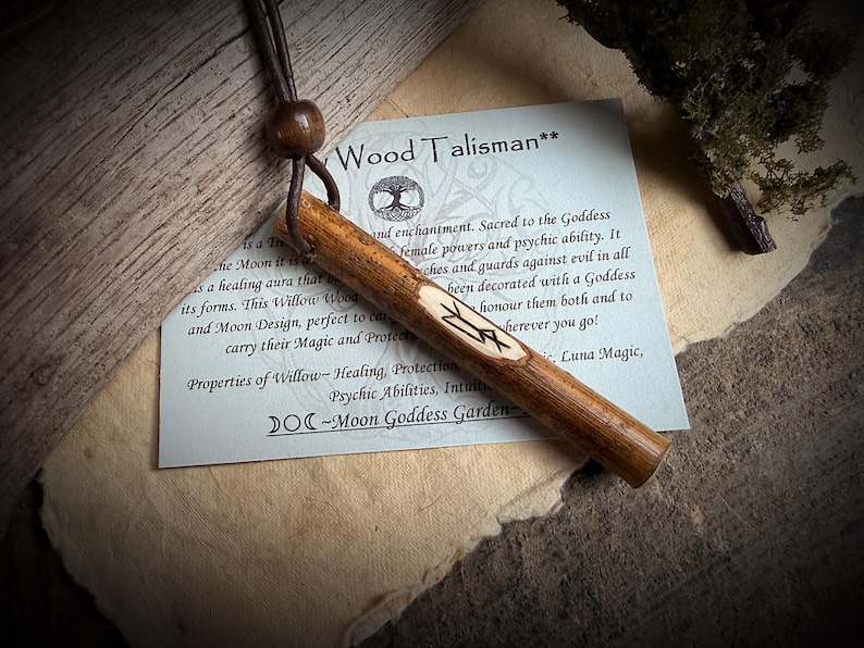 K&ouml;nnte beinhalten: Ein Holz-Talisman mit einer Lederschnur und einer Holzperle. Der Talisman ist zylindrisch, mit einem geschnitzten Symbol. Eine Karte mit Text &uuml;ber die Eigenschaften des Talismans liegt daneben. Der Text auf der Karte lautet "Wood Talisman".