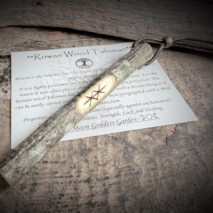 May include: A Rowan Wood Talisman, a rustic wooden pendant with a carved rune design, is displayed on a textured surface. The pendant is attached to a brown leather cord. A card with text about the talisman is visible.