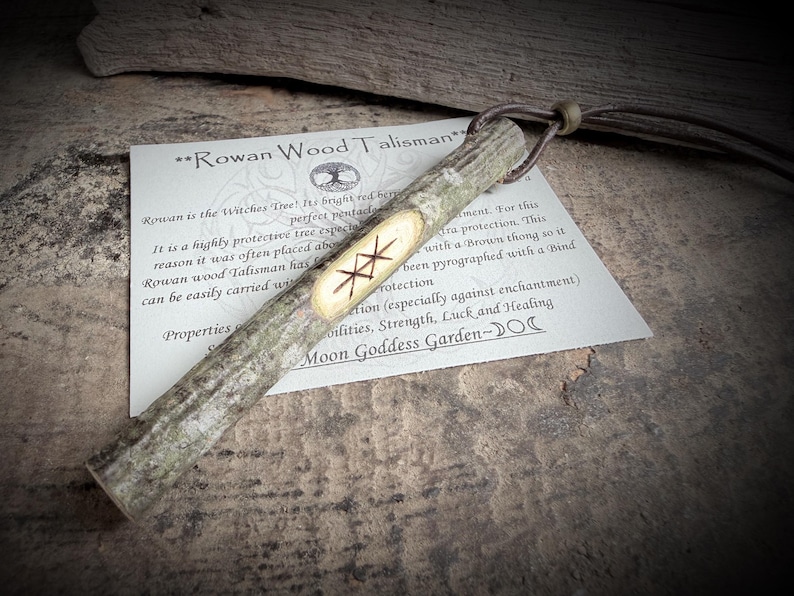 May include: A Rowan Wood Talisman, a rustic wooden pendant with a carved rune design, is displayed on a textured surface. The pendant is attached to a brown leather cord. A card with text about the talisman is visible.